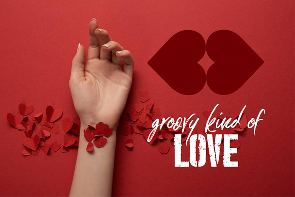cropped view of female hand with paper cut decorative hearts on red background with "groovy kind of love" lettering  - Foto, imagen