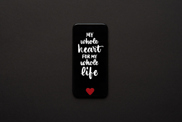 κάτοψη του smartphone με γράμματα «όλη την καρδιά μου για όλη μου τη ζωή» και μικρή κόκκινη καρδιά σημάδι σε σκούρο φόντο - Φωτογραφία, εικόνα