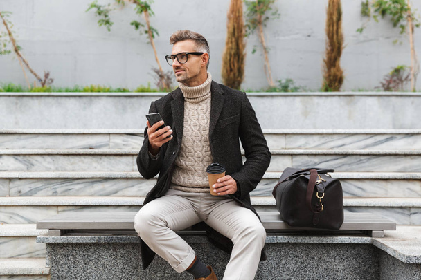 Handsome man wearing jacket holding mobile phone, drinking takeaway coffee while sitting outdoors - Photo, image