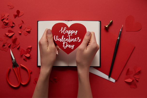 cropped view of woman holding paper cut heart sign with "Happy valentines day" lettering on red background - Photo, image