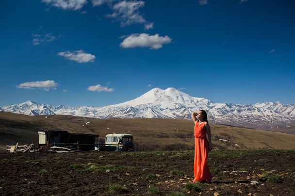 portrait of a full-length slender brunette girl in a long red dress in the field in front of high mountains - Foto, afbeelding