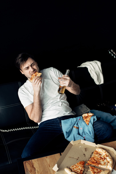 μεθυσμένος άντρας τρώει νόστιμο πίτσα και κρατώντας το μπουκάλι στο βρώμικο σαλόνι - Φωτογραφία, εικόνα