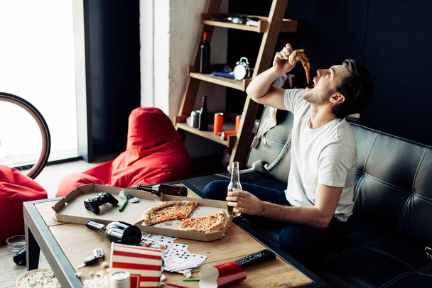 man eating pizza while holding bottle in messy living room - Photo, image