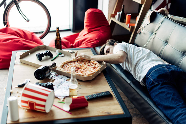 Betrunkener liegt nach Party auf Sofa neben Couchtisch in schmuddeliger Wohnung - Foto, Bild