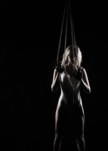 Beautiful slender blonde girl with big breast and nipples appearing through clothes, wearing a white bodisuit, sensually plays with the ropes on black. Artistic noir silhouette photo. Copy space. - Фото, зображення