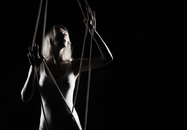 Beautiful slender blonde girl with big breast and nipples appearing through clothes, wearing a white bodisuit, sensually plays with the ropes on black. Artistic noir silhouette photo. Copy space. - Фото, зображення