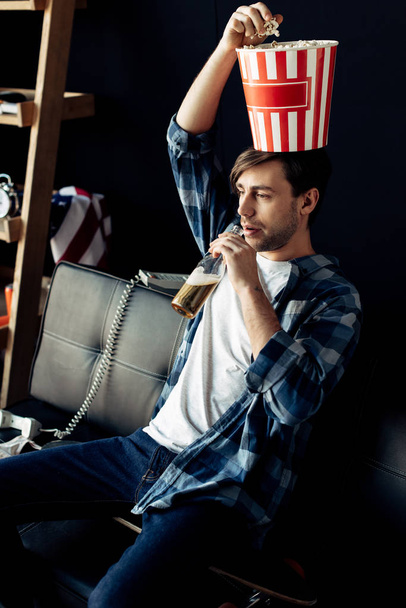 handsome man holding popcorn box on head drinking beer from bottle at home - Foto, imagen