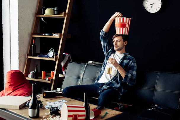 man holding popcorn box on head and sitting on sofa in messy living room - Foto, Bild