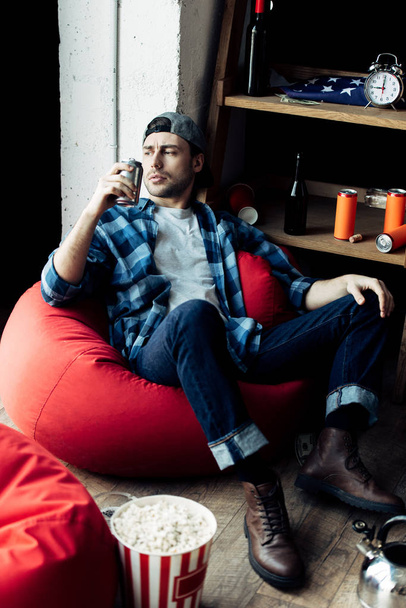 όμορφος άντρας πίνει μπύρα από μπορεί και να κάθεται στην τσάντα φασολιών στο σπίτι  - Φωτογραφία, εικόνα