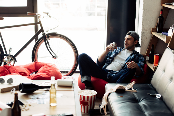 Selektivní fokus člověka pití piva z can a sedí na bean bag nepořádek doma  - Fotografie, Obrázek