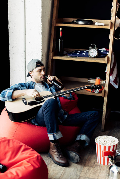 humalassa oleva mies juo olutta ja pitelee akustista kitaraa kotona
  - Valokuva, kuva