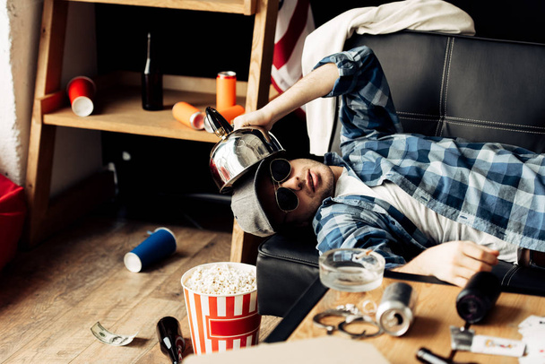 hombre guapo sosteniendo la tetera cerca de la cabeza mientras está acostado en el sofá en la sala de estar desordenado
  - Foto, Imagen