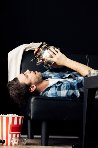 przystojny mężczyzna picia z czajniczek, leżąc na kanapie w brudny pokój dzienny  - Zdjęcie, obraz