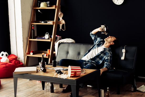 drunk man drinking from teapot while sitting on sofa in messy living room  - Фото, изображение