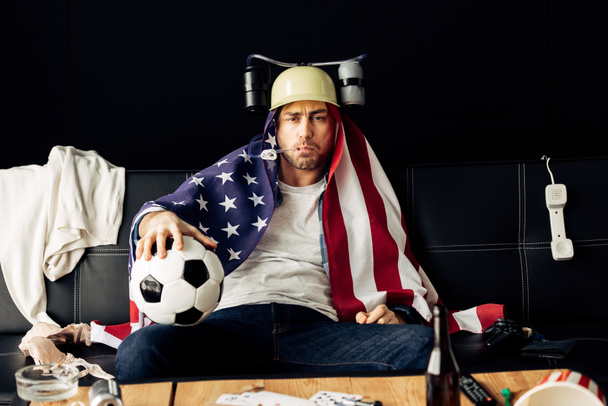 man wearing beer helmet drinking and holding football while sitting with american flag on shoulders - Photo, Image