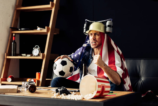 man wearing beer helmet drinking and holding ball while sitting with american flag on shoulders and watching game - Photo, Image