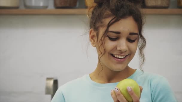 Beautiful young slender brunette with green apple in one hand and croissant in the other hand on background of vegetables on kitchen table making a difficult choice between healthy and harmful food. - Materiaali, video