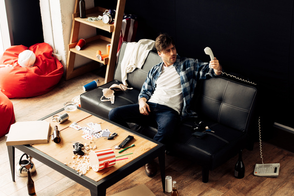 drunk man looking at retro phone and sitting on sofa in messy home - Photo, Image