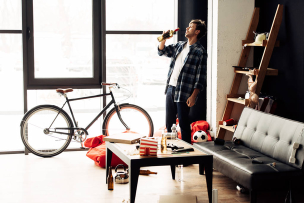 homem bebendo vinho de garrafa na sala de estar bagunçada após a festa
 - Foto, Imagem