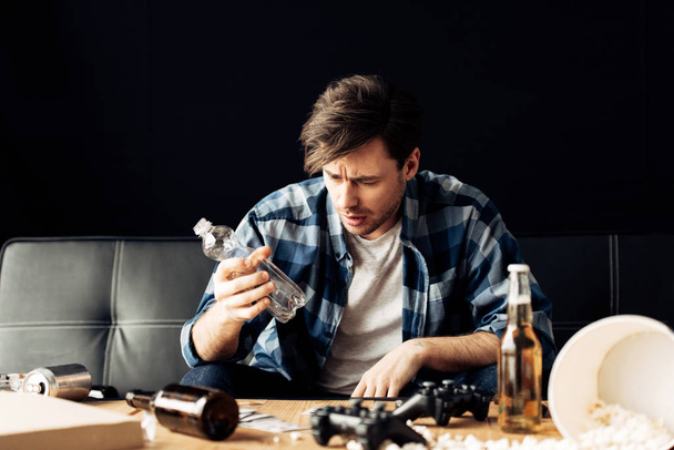 handsome man suffering from hangover looking at empty bottle while sitting on sofa - Φωτογραφία, εικόνα