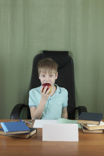 Умный школьник сидит за столом с большим количеством книг и красным яблоком. Концепция образования
.  - Фото, изображение