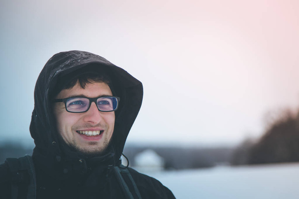Νέος, χαμογελαστός άνθρωπος σε ένα μαύρο σακάκι με κουκούλα, χειμώνα εκτός - Φωτογραφία, εικόνα