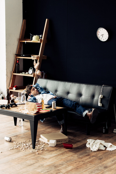 zmęczony człowiek leżący na kanapie po imprezie w brudny pokój dzienny  - Zdjęcie, obraz