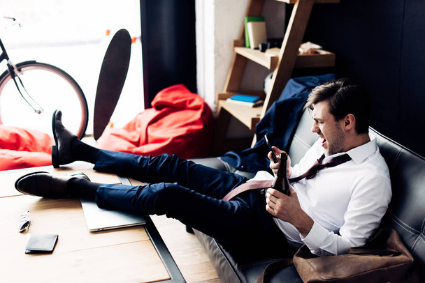 homem de terno segurando garrafa e gritando com smartphone enquanto sentado no sofá após a festa
 - Foto, Imagem