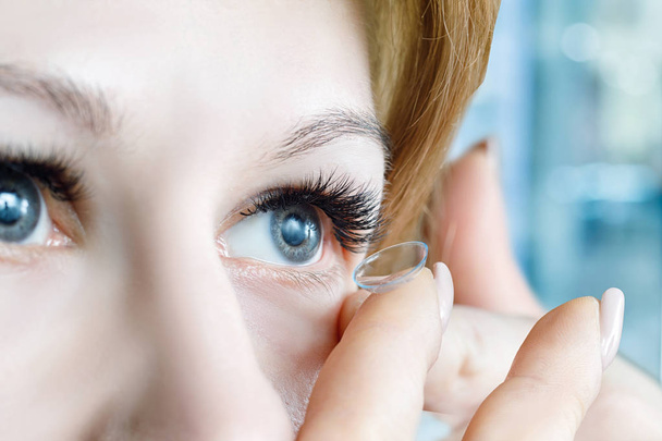 Een close-up van een jonge vrouw die een transparante contactlens invoegen in haar oog bij wazig ziekenhuis kamer achtergrond. Het concept van oogheelkundige slechte zicht correctie. - Foto, afbeelding