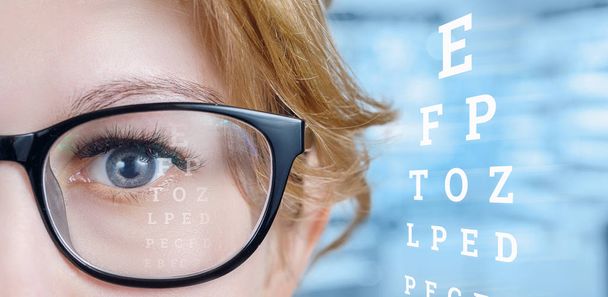 Closeup ženské šedých očí s brýlemi při pohledu rovně a tabulku test pohled na pozadí rozmazané ordinace. - Fotografie, Obrázek