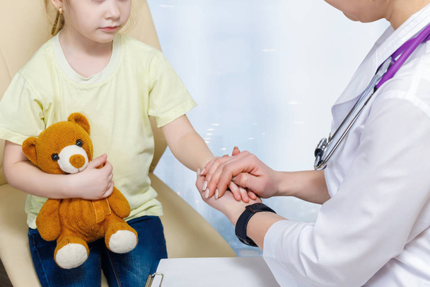 Un primer plano de la pediatra que sostiene una mano de un niño pequeño sentado con un oso de juguete con gesto protector para apoyar antes del tratamiento en un fondo borroso
. - Foto, imagen