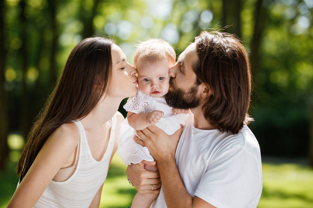 Romantic family on the open air. Young dark-haired woman and her husband are kissing their charming little daughter. - Zdjęcie, obraz