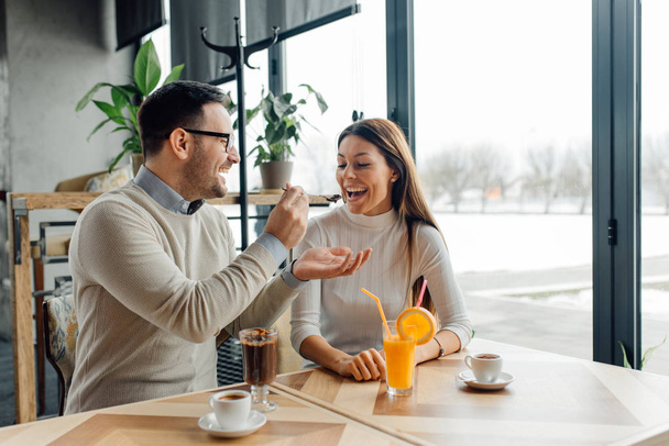 Handsome boyfriend feeding smiling girlfriend with tasty dessert during date in cafe - Foto, imagen