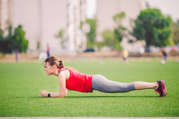 Theme sport and health. Young Caucasian woman doing warm-up, warming up muscles, training abdominal muscles. Losing belly. Abdominal plank exercise on green grass in stadium summer artificial turf - Photo, Image