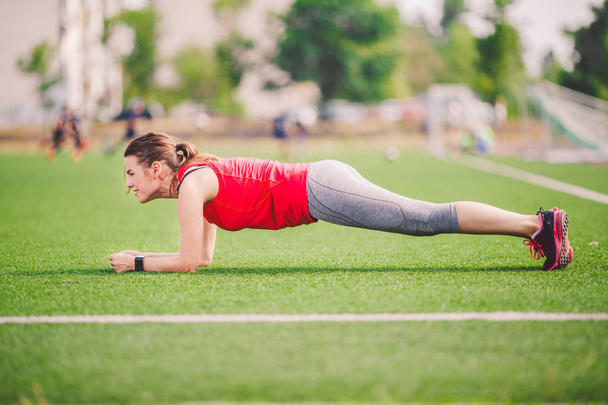 Theme sport and health. Young Caucasian woman doing warm-up, warming up muscles, training abdominal muscles. Losing belly. Abdominal plank exercise on green grass in stadium summer artificial turf - Fotó, kép