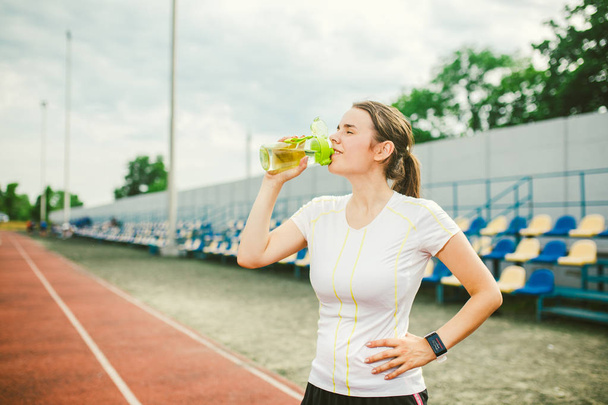 Thema ist Sport und Gesundheit. schöne junge kaukasische Frau mit großen Brüsten Athlet Läufer steht auf Laufstadion ruhen, Laufstrecke mit Flasche in den Händen trinken Wasser in kurzen Shorts - Foto, Bild