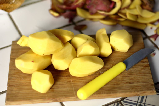 A peeled raw potato, a vegetarian ingredient for cooking meals, on a wooden board next to a potato peel and a knife for peeling potatoes in the kitchen. Dinner preparation process - Photo, Image