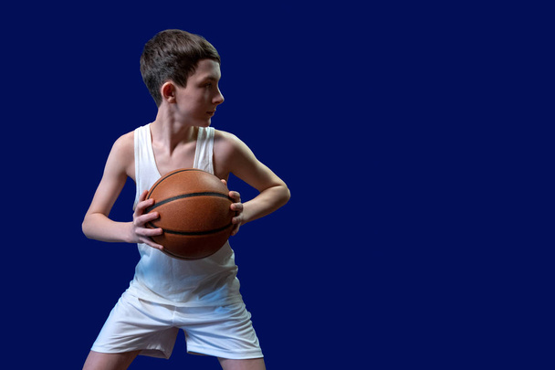 Um adolescente com uma camiseta branca e shorts esportivos segura uma bola de basquete e olha para o lado. Espaço para cópia. Em branco para cartaz desportivo. Isolado em fundo azul
. - Foto, Imagem