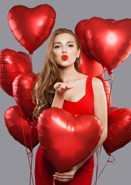 Νεαρή όμορφη γυναίκα φυσώντας φιλί και κρατώντας κόκκινα μπαλόνια. Μοντέλο κορίτσι με κόκκινα χείλη μακιγιάζ φοράει κόκκινο φόρεμα σε φόντο Εορτασμός Κόμμα διακοπών. Έννοια αγάπης, δώρο και ημέρα του Αγίου Βαλεντίνου - Φωτογραφία, εικόνα