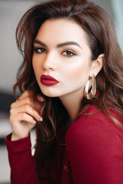 Portrait of gorgeous young brunette woman with make up, red lips, golden earrings wearing red blouse. She is looking at camera. - Photo, Image