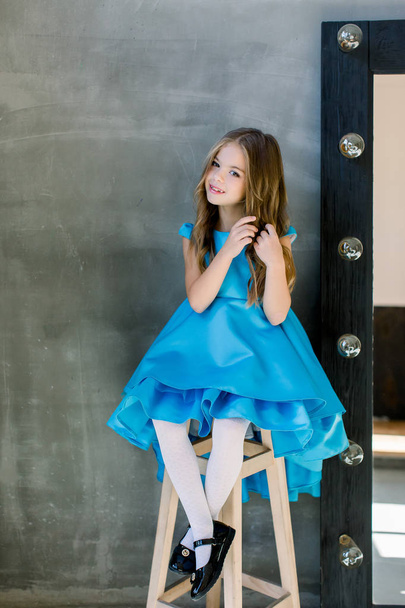 portrait of beautiful delighted little lady in a blue dress is sitting on the stool and touches the hair - Foto, Imagem
