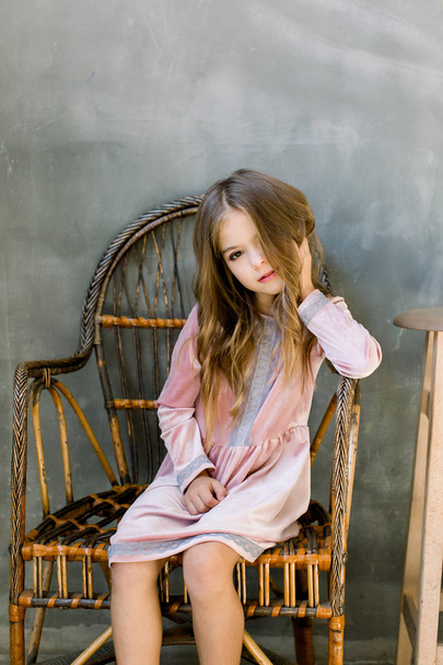 Roztomilá holčička 5-6 let staré, stylové růžové šaty sedí na pozadí dřevěná židle overgrey. Při pohledu na fotoaparát. Oslava narozenin. Oslava. - Fotografie, Obrázek