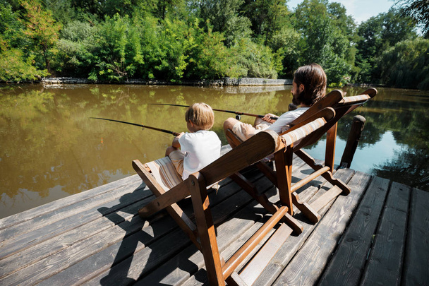 Young dark-haired father and his little son are sitting in recliners on the wooden pier with fishing rods and fishing. - Foto, Bild