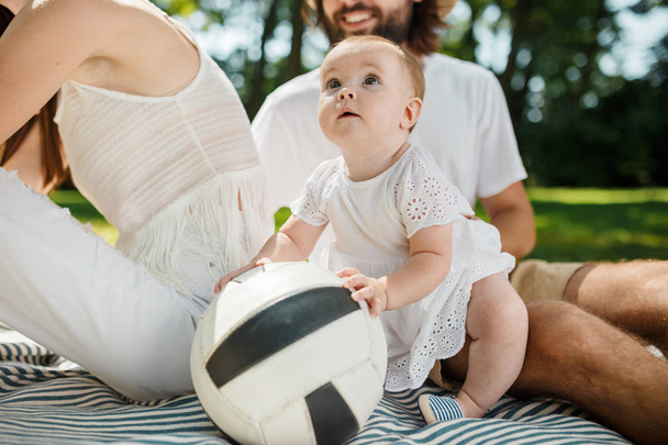 Adorable little girl in white dress sits next to parents on a blanket on the lawn and touches a football ball. - Foto, imagen