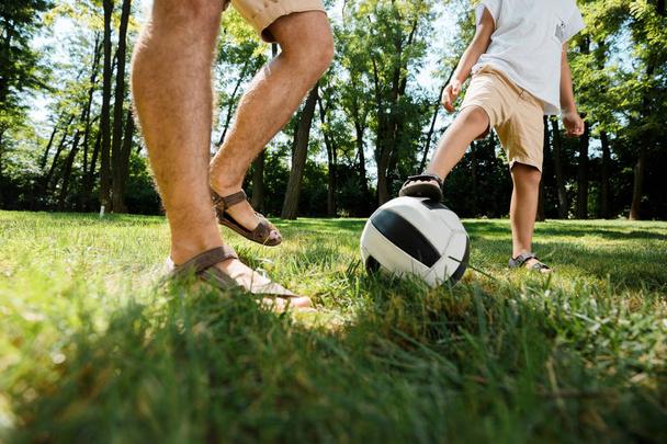 Handsome father and his little son dressed in the white t-shirts are playing football on a lawn on a warm day. - Fotó, kép