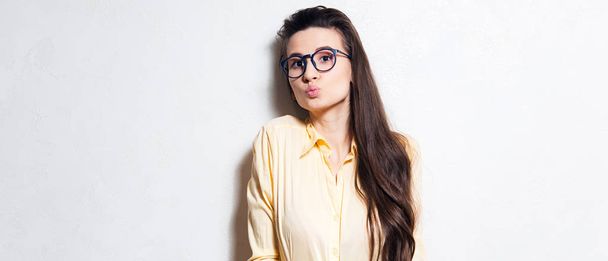 Young cheerful girl sending blowing kiss with pout lips. Wearing blue glasses and yellow shirt. Over white background - Photo, Image