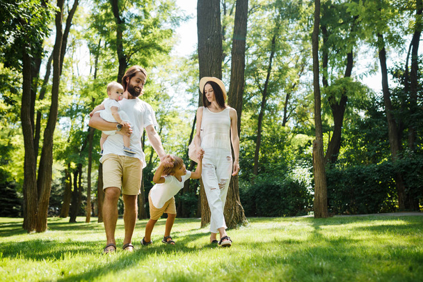 Dia de verão ensolarado. Jovem família feliz vestida com as roupas casuais brancas caminha no parque
. - Foto, Imagem