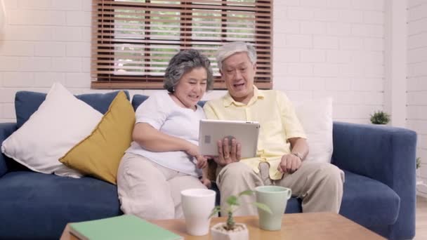 Asian elderly couple using tablet watching TV in living room at home, couple enjoy love moment while lying on sofa when relaxed at home. Enjoying time lifestyle senior family at home concept. - Footage, Video