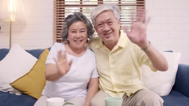 Ázsiai idős pár használja okostelefon video konferencia unokája, miközben feküdt kanapén a nappaliban otthon. Élvezi az időskori életstílust otthon. Portré a kamerába néz. - Felvétel, videó