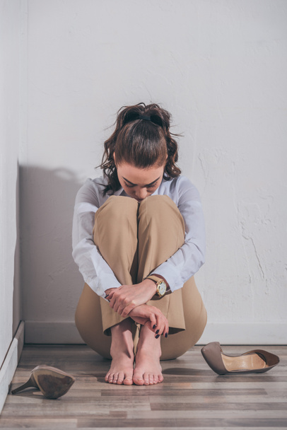 mujer triste en blusa blanca y pantalones beige sentado en el suelo, abrazando las rodillas cerca de la pared en casa, concepto de trastorno de duelo
 - Foto, Imagen